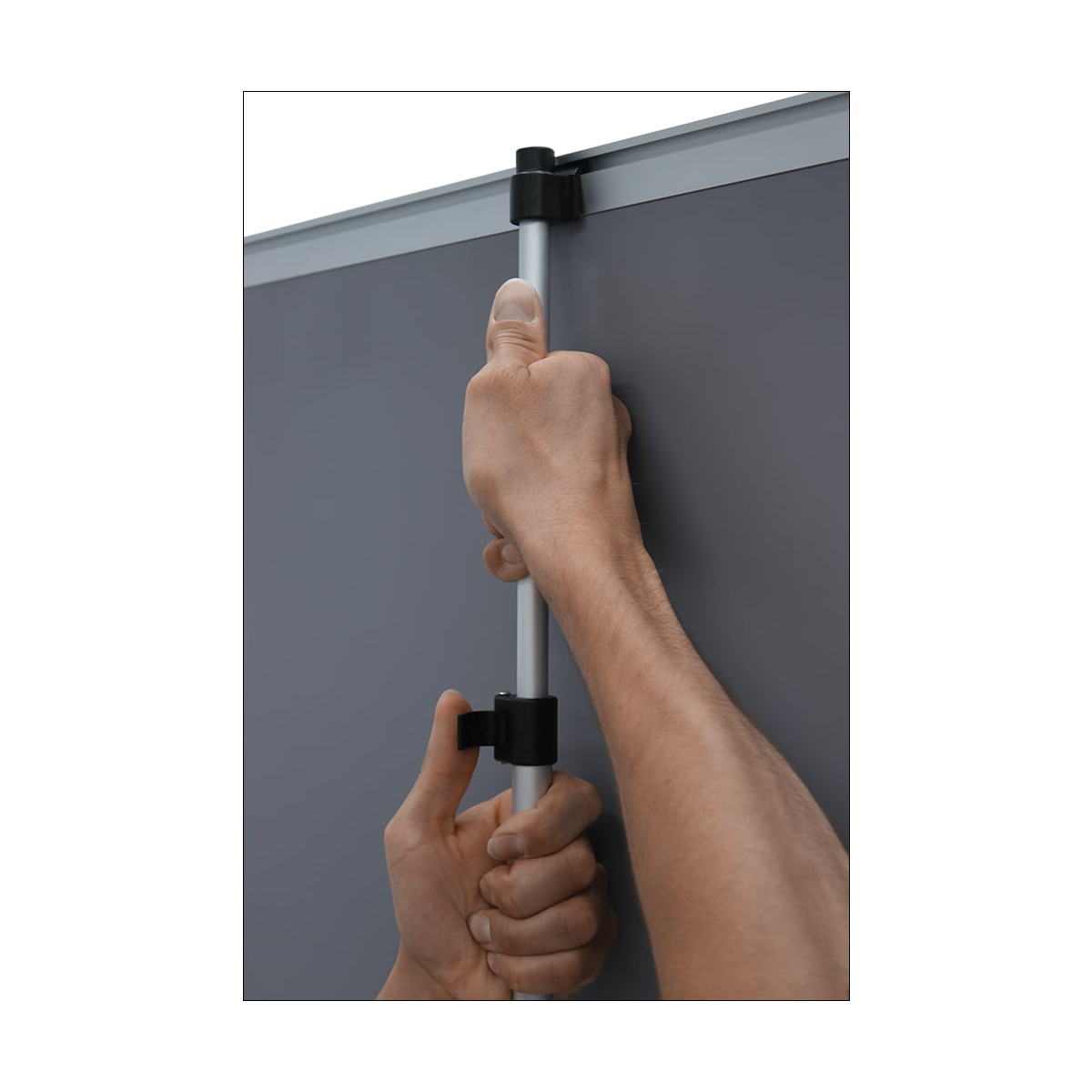 A close-up of a person's hands adjusting a silver metal pole with black plastic clamps against a dark gray screen or panel.